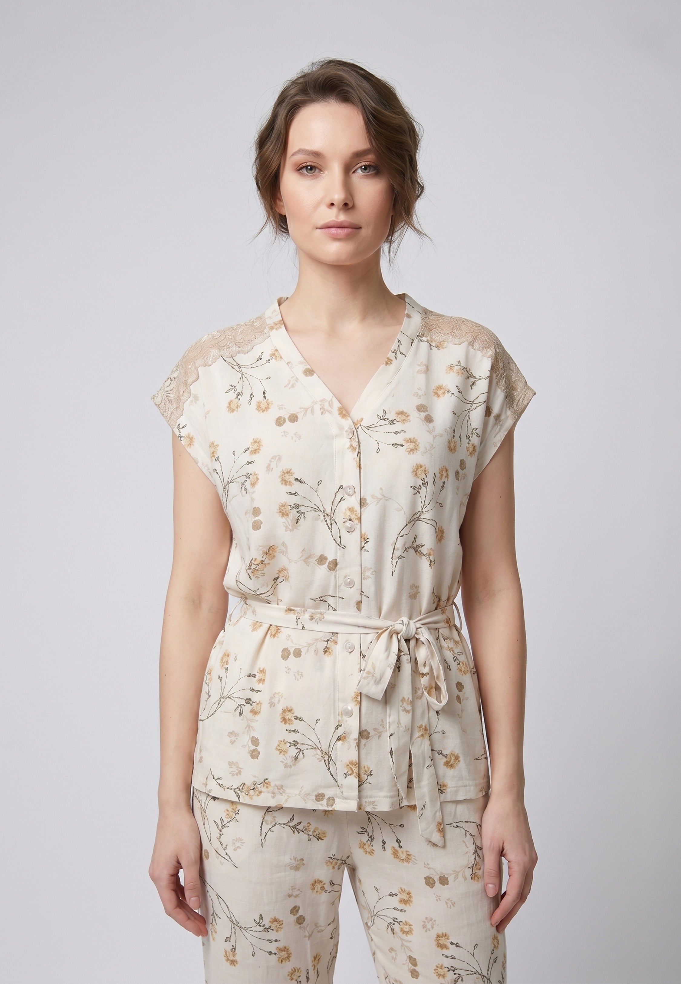 Femme portant un pyjama deux pièces blouse boutonnée col V sans manches empiècement dentelle épaules ceinture nœud imprimé fleuri beige et doré sur fond crème et pantalon droit assorti Rosabella Soft Bloom