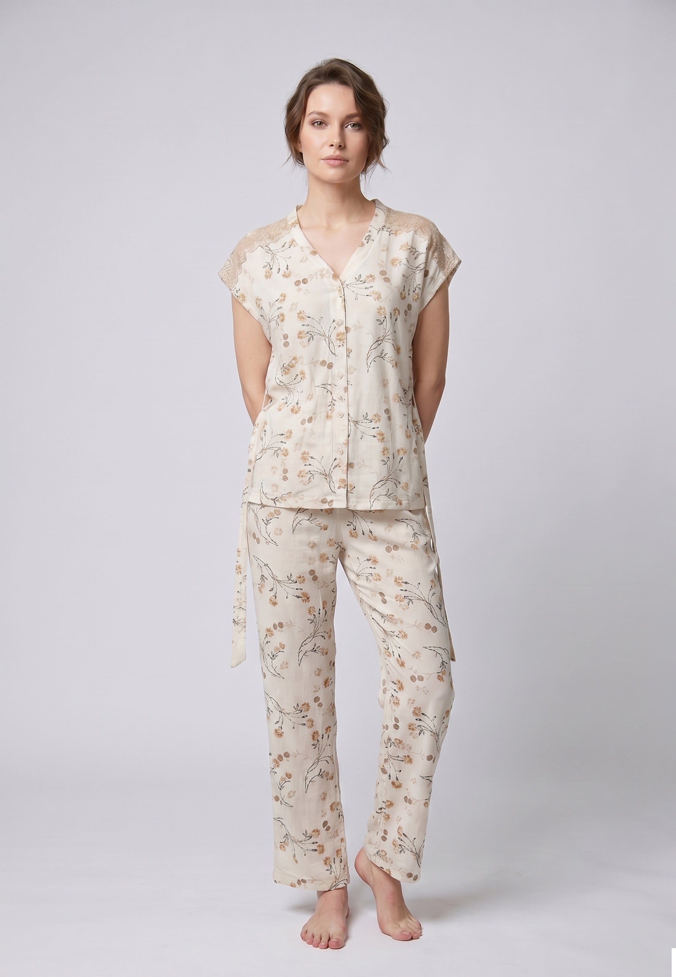 Femme portant un pyjama deux pièces blouse boutonnée col V sans manches empiècement dentelle épaules ceinture nœud imprimé fleuri beige et doré sur fond crème et pantalon droit assorti Rosabella Soft Bloom