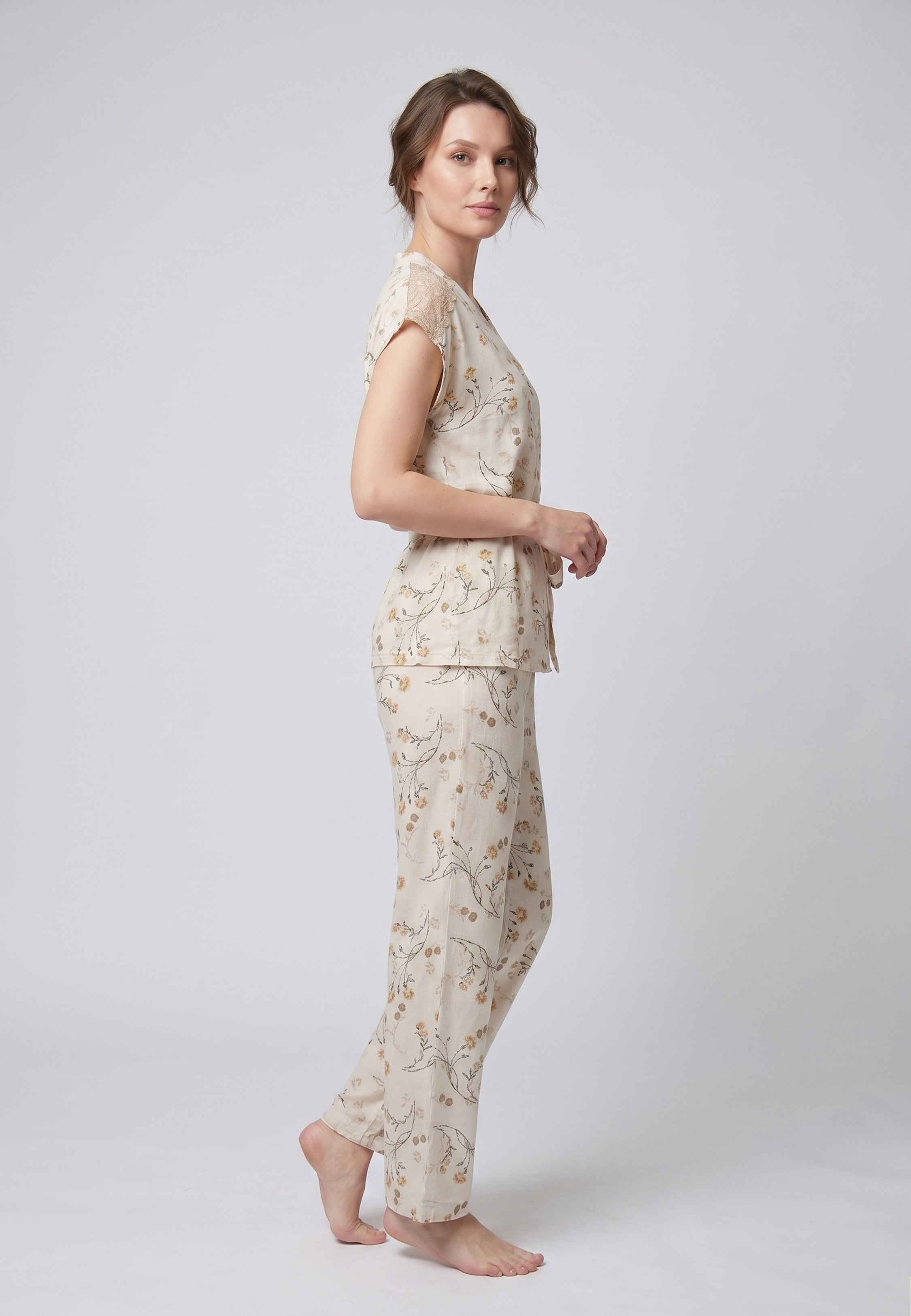 Femme portant un pyjama deux pièces blouse boutonnée col V sans manches empiècement dentelle épaules ceinture nœud imprimé fleuri beige et doré sur fond crème et pantalon droit assorti Rosabella Soft Bloom