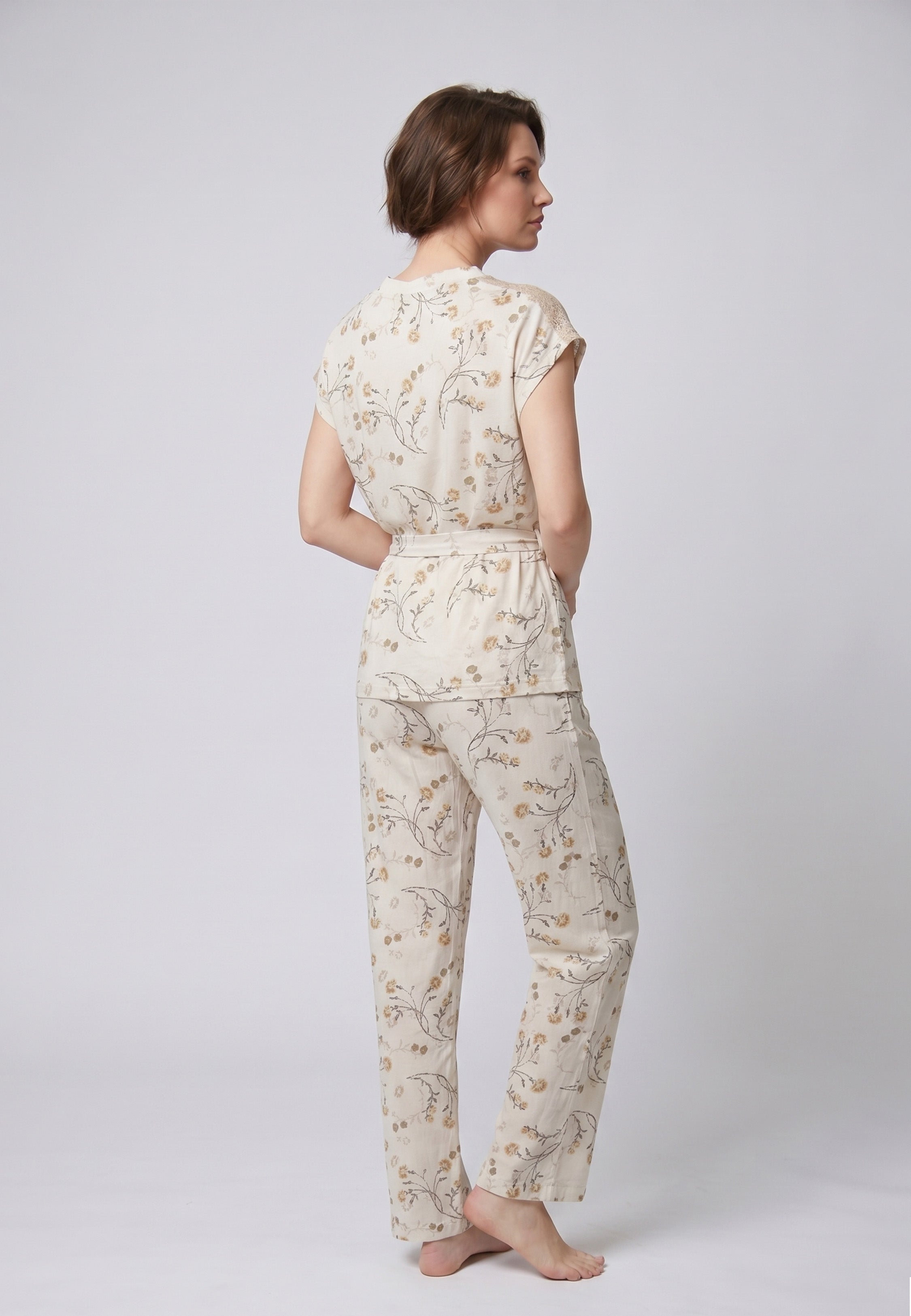 Femme portant un pyjama deux pièces blouse boutonnée col V sans manches empiècement dentelle épaules ceinture nœud imprimé fleuri beige et doré sur fond crème et pantalon droit assorti Rosabella Soft Bloom
