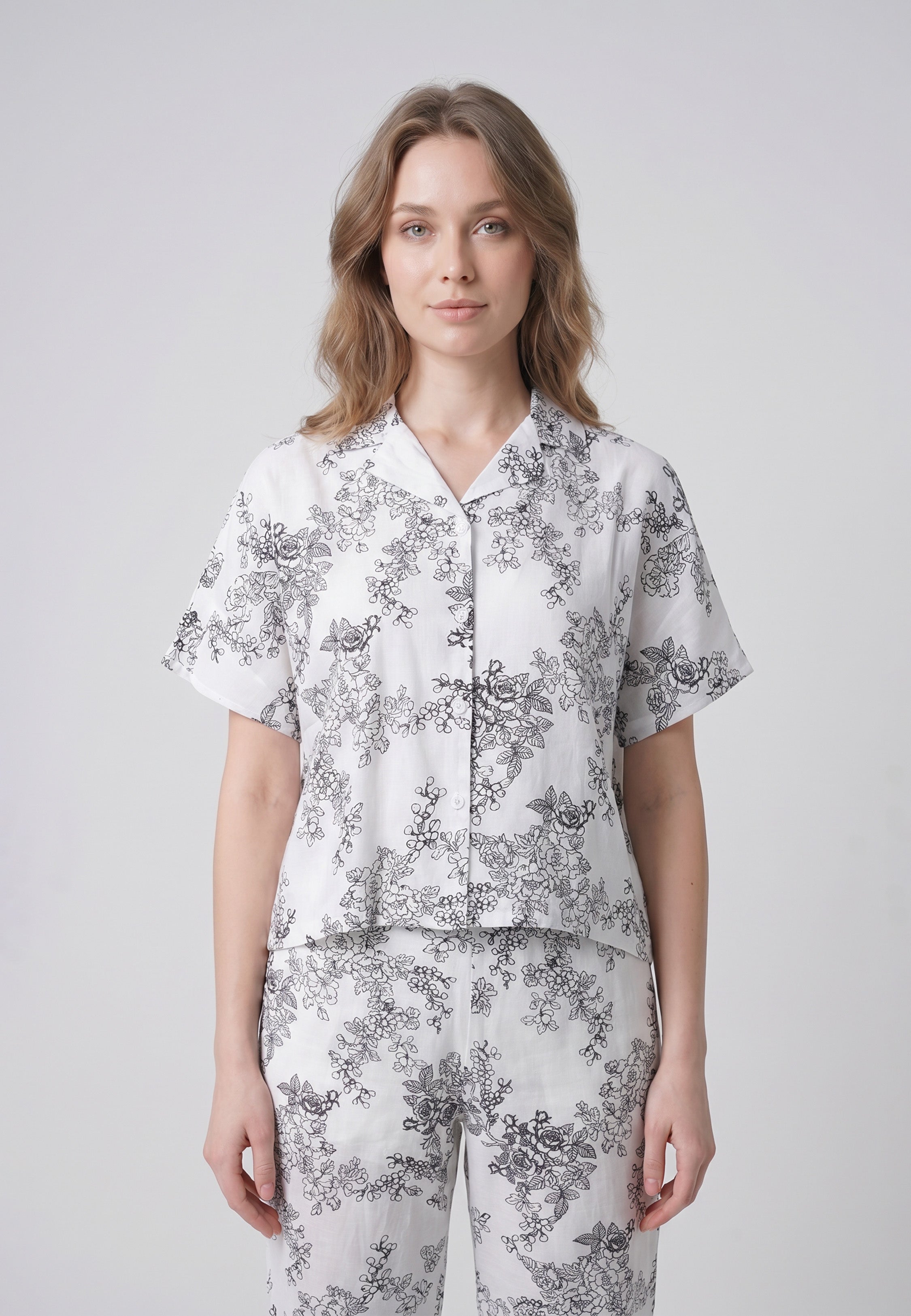 Femme portant un pyjama deux pièces chemise boutonnée col pyjama manches courtes imprimé botanique noir fond crème et pantalon droit assorti Rosabella Calm Evenings
